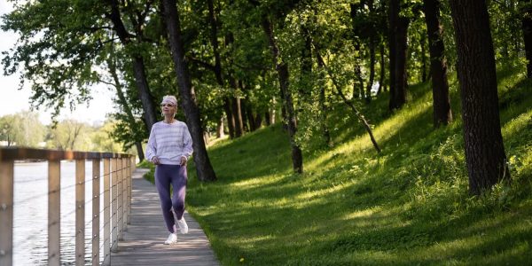 Elderly woman running along a riverside path in a green park, embracing fitness and nature.